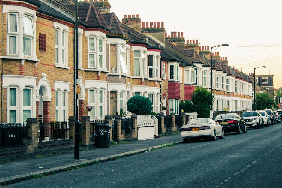 Photograph of a residential street during late afternoon or early evening, featuring a row of traditional brick terraced houses with white window frames and bay windows, some with small front gardens 