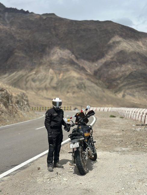 A black and silver Himalayan motorcycle parked outdoors on a sandy surface with a mountainous landscape in the background under partly cloudy skies. The motorcycle is positioned in profile, showing it