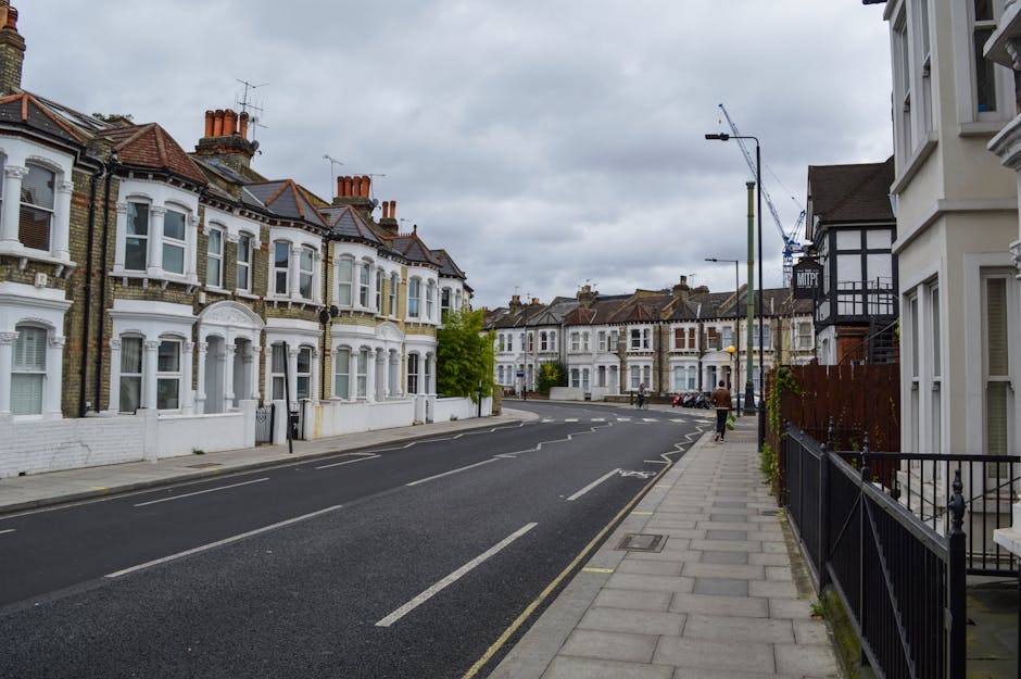 A wide view of a residential street in Enfield Highway on a cloudy day, featuring Victorian-style terraced houses with white facades and red brick chimneys on the left side, and modern white buildings with bay windows on the right side. The street has a paved sidewalk with black metal railings, and a few pedestrians walking along the pavement. Parked cars are visible along the curb, with some parked on the street and others on the driveway of the houses. Streetlights and a crane are visible in the distance, indicating nearby construction. The scene is calm with minimal activity, capturing the typical appearance of a suburban house area suitable for home relocation or furniture transport services. Man with Van Enfield Highway occasionally provides such house removals and packing services in this area, as reflected by the environment and typical relocation setting.