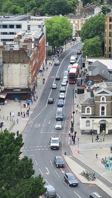 An aerial view of a busy street near Enfield Town station area in Enfield Highway, showing parked and moving cars along a two-lane road with a dedicated left-turn lane, bordered by mature green trees and various brick and stone buildings, including commercial premises and residential houses. Pedestrians walk along the wide pavements, and bicycles are parked near the sidewalk. The scene captures a typical urban environment during daytime, with vehicles waiting for the traffic lights to change. This setting is relevant for house removals and furniture transport services provided by Man with Van Enfield Highway, emphasizing the logistical context of loading or unloading belongings during a home relocation or moving process.