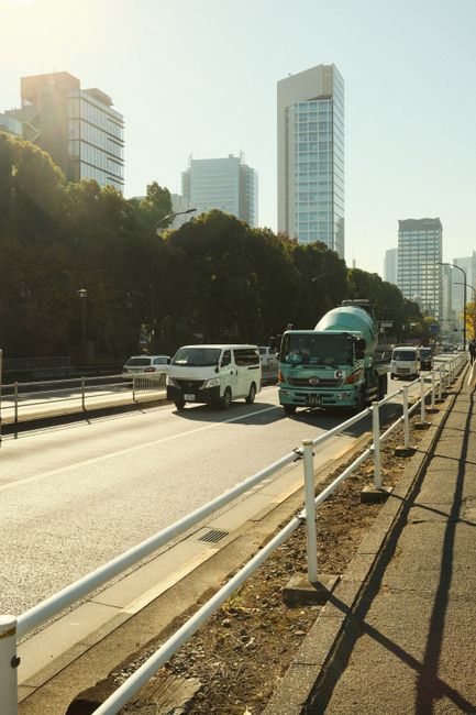 A city street scene during daytime featuring a multi-lane road with several vehicles, including a white van and a turquoise truck, moving along the lanes. The vehicles are in the process of being loaded or unloaded as part of a house or office relocation, with visible materials such as cardboard boxes and wrapped furniture inside the truck. The scene is set beside a sidewalk with a white metal railing, which separates the pedestrian pathway from the roadway. In the background, tall modern office buildings and skyscrapers made of glass and steel rise against a bright sky, with sunlight casting shadows across the scene. The setting suggests a busy urban environment suitable for professional removals or moving services, with a focus on transportation and logistics involved in furniture transport and home or office relocation, as provided by Man with Van Enfield Highway.