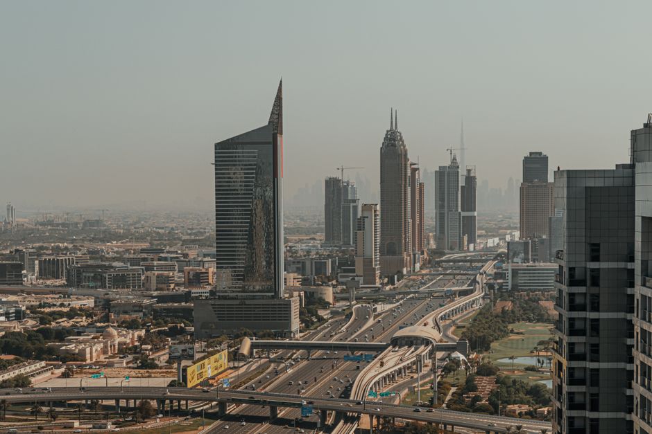 An aerial view of a cityscape featuring a modern, angular glass skyscraper and several other high-rise buildings with various architectural styles, including a historical gothic-style tower with spires. The scene shows a busy multilane highway with overpasses and ramps, with several vehicles including cars and trucks, some parked near the buildings. In the foreground, there is a paved area adjacent to the buildings, possibly a loading or parking zone, with visible shadows suggesting sunlight from the left. This urban environment illustrates a city centre suitable for professional removals and furniture transport services, such as those offered by Man with Van Enfield Highway, involved in house and office relocations, including packing, loading, and logistics coordination in high-density areas.