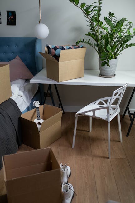 Inside a room during a house move, there are three cardboard boxes stacked on top of each other near the wall, with the largest box at the bottom and the smallest on top. The boxes are sealed with pac