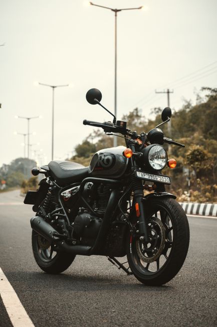 A black motorcycle with a streamlined design is parked on the side of a rural road, featuring dual rearview mirrors, a round headlamp, and an orange indicator light on each side. The motorcycle has a matte black engine, exhaust pipe, and wheels, with a grey seat extending over the frame. In the background, there are tall, evenly spaced streetlights lining the road, which curves slightly to the left. The sky appears overcast with a muted light, and distant hills covered with trees are visible on the horizon. The scene suggests a quiet area suitable for home relocation or vehicle transportation, illustrating the kind of transport used in moving and logistics services. The company Man with Van Enfield Highway specializes in removals and furniture transport, supporting home relocation and packing and moving processes. The motorcycle is parked close to the pavement, captured in a still position, emphasizing the importance of secure vehicle storage during a move.