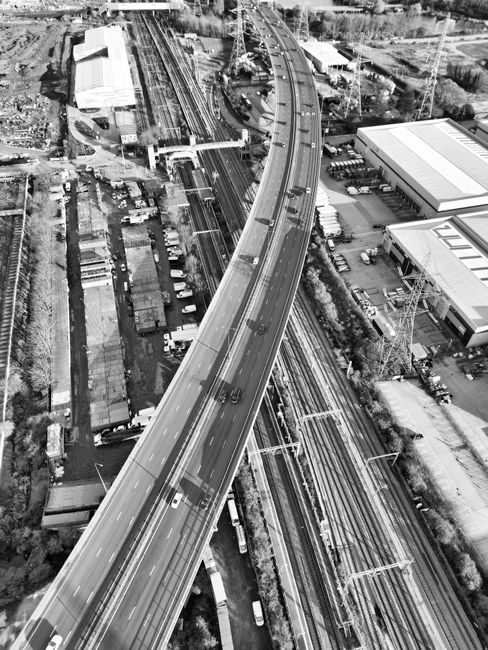 A black-and-white aerial photograph showing a section of urban infrastructure with a prominent elevated motorway curving through the scene. Below the motorway, multiple sets of railway tracks run parallel, with some trains and railcars visible alongside station platforms. Adjacent to the railway, there are industrial and commercial buildings, some with flat roofs and large windows. The area includes parking lots filled with vehicles, alongside narrow roads and pedestrian pathways. Surrounding greenery and trees are present around the perimeter of the industrial zone. The lighting suggests daytime, with shadows cast by the structures, and the overall view captures the dense layout of transportation and industrial facilities in the area. This type of scene relates to logistics and transport services involved in house removals and furniture transport, as provided by Man with Van Enfield Highway.